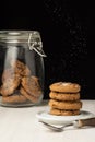 View of stack of chocolate cookies on plate, with falling sugar, spoon and glass jar with cookies, selective focus, on white woode Royalty Free Stock Photo
