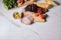 View of tasty ham on cutting board with parsley, cherry tomatoes, olives and baguette on white marble surface
