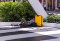 View of tired man with yellow bag sleeping on floor near green bushes on walking street. Miami. USA