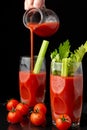 View of two glasses with Bloody Mary cocktail with celery, woman`s hand serving from a jug, and cherry tomatoes, on black backgro Royalty Free Stock Photo