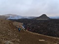 View of volcanic eruption at Fagradalsfjall mountain with people passing by a black colored, glowing lava field with smoke.