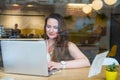 View through the window with city reflection of smiling businesswoman in headset working on laptop sitting in cafe Royalty Free Stock Photo