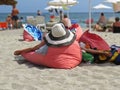 View at Woman from Back Lying on Red Pillow Cushion and Relaxing on Ocean Sand Beach Wearing Straw Hat During Summer Vacation Royalty Free Stock Photo