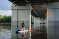 View on woman which stands on sup board and swim on the river under bridge.