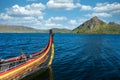 Viking warrior ancient wooden long ship on sea with blue cloudy sky and green mountain in the background.