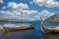 Viking warrior ancient wooden long ships in harbour with blue cloudy sky and green mountain in the background.