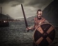 Viking warrior in the attack, standing along the shore with Drakkar and mountains on the background.