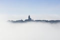 Village covered in fog with some houses and the church tower protruding above the clouds.