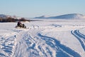 The snowmobile with fishermen is riding along the winter lake against the backdrop of the mountains.