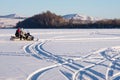 The snowmobile with unidentified fishermen is riding along the winter lake against a background of a hill with birches.