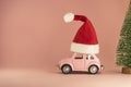 Pink retro toy model car with small red Christmas Santa Claus hat and little Xmas tree on pastel pink background