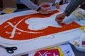 People carefully glue seeds of lentils and rice to a drawing of a city emblem on a big white wooden plate
