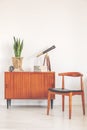 Vintage cupboard with plant, books, telescope and clock next to stylish wooden chair in white and bright interior with copy space