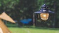 Vintage hanging black camping lantern with blurred background of field tents in camping area at natural parkland