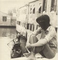 Girl sitting on the side of a bridge in Venice watching a gondola pass, in the 60s