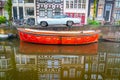 Vintage orange color canal boat moored alongside with white American classic car parked on road in front of typical Dutch canal Royalty Free Stock Photo