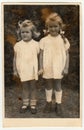 Vintage photo shows two small girls - siblings. They wear a white dresses. Retro black and white photography. Circa