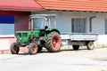 Vintage retro old green tractor with open cabin and attached strong light grey trailer parked next to abandoned industrial
