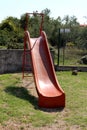 Vintage retro old red plastic playground slide with metal stairs surrounded with grass and stone wall with metal fence in Royalty Free Stock Photo