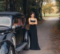 Vintage style photo. Elegant retro woman in black evening long dress posing on road, autumn nature background green