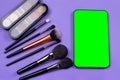 Violet background with decorative cosmetic palette and different brushes for makeup. Smartphone with green screen near make-up set Royalty Free Stock Photo