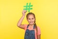 Viral web content, internet forum. Portrait of happy little girl with braid in denim overalls holding hashtag symbol