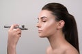 Visagist making makeup for model with aerograph, isolated on grey. Background with hand holding aerograph. Visagist using aerograp