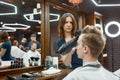 Visiting beauty salon. Young guy sitting in barbershop chair while professional focused barber girl drying his hair Royalty Free Stock Photo