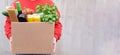 A volunteer, a food delivery man in a red sweater or jacket, is holding a box of groceries, greens, pasta, pasta, wine, vegetables