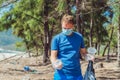 Volunteer man in face mask pick up garbage pollute beach near sea, hold look at discarded disposable plastic cups