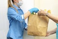 Volunteer young blonde female in blue uniform and medical mask and gloves handing an senior woman a paper bag with food. Donation Royalty Free Stock Photo