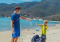 Volunteers paradise beach sand lazur sea. Man, boy pick up garbage into black bag. Son refuse to wear blue face mask