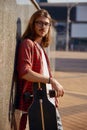 Waist portrait of young hipster man standing with longboard leaning back to wall