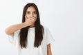 Waist-uo shot of concerned shocked female student in white t-shirt, covering mouth with palm and staring at camera with