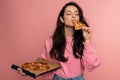 Hungry dark-haired young female eating an appetizing Italian food on the pink background during the studio photo shoot