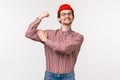 Waist-up portrait of funny adult slim guy in red beanie and glasses acting cool and tough, flex biceps showing how