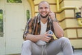 Waist up portrait of young good looking bold bearded guy is sitting outdoors on stairs in front of his house holding cellphone in Royalty Free Stock Photo