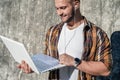 Waist up portrait of young good looking bold bearded hipster standing outdoors against grey modern loft wall with laptop and Royalty Free Stock Photo