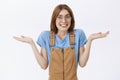 Waist-up shot of awkward cute charismatic young woman in brown overalls shrugging with hands near shoulders smiling with