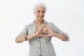 Waist-up shot of charming caring granny with grey hair in shirt showing heart sign over chest and smiling broadly loving