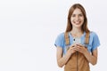 Waist-up shot of charming good-looking young female smartphone user in trendy overalls smiling joyfully with friendly Royalty Free Stock Photo