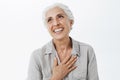 Waist-up shot of happy and delighted grateful charming old lady with white hair holding palm on chest in thankful