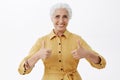 Waist-up shot of pelased kind and nice elegant old lady in trendy yellow coat showing thumbs up approving great idea or