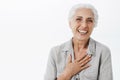 Waist-up shot of pleased grateful charming granny in shirt holding palm on chest and smiling broadly at camera being