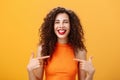 Waist-up shot of proud and happy outgoing charming woman with curly hair and red lipstick pointing at herself delighted