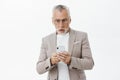 Waist-up shot of shocked mature male digital nomad in suit and glasses with grey hair and beard holding smartphone