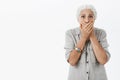 Waist-up shot of shocked and stunned elderly woman in casual shirt with grey hair gasping covering mouth with both palms