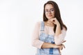 Waist-up shot of smart and creative female artist in glasses and overalls holding hand on jawline and smiling pleased