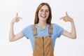 Waist-up shot of smart self-assured european woman in glasses and overalls pointing at herself with proud confident