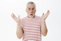 Waist-up shot of surprised handsome senior man with grey beard in prescribed glasses and striped t-shirt raising palms Royalty Free Stock Photo